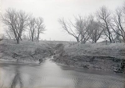 black and white photo of a small stream with dirt banks, long grass, and trees without leaves
