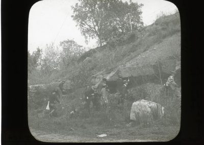 Black and white photograph of a hill with rock ledge with several men at the base of rock ledge
