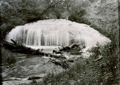 black and white photo of waterfall amond shore bluffs and tree limbs in stream