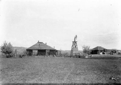black and white photo og sod house and buildings, windmill, and wagons on a farm in open fields